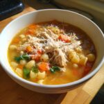 A close-up of a white bowl filled with hot Sicilian chicken soup, topped with shredded chicken and grated Parmesan cheese.
