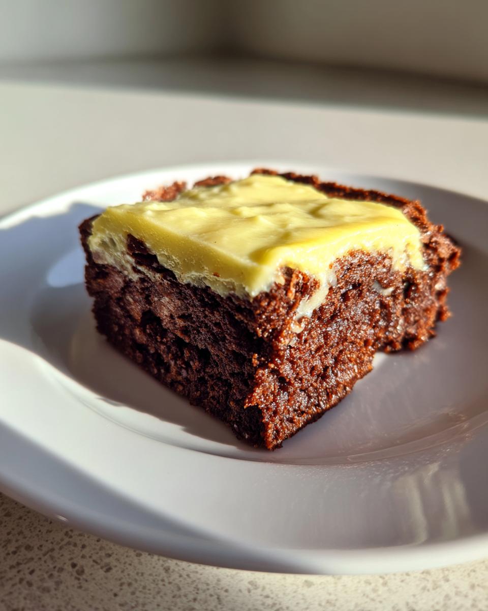 A close-up of a rich, dark chocolate gingerbread brownies square topped with a thick layer of pale yellow cream cheese frosting.