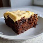 A close-up of a rich, dark slice of gingerbread brownies topped with thick, pale yellow frosting.