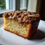 A close-up shot of a moist slice of coffee cake featuring a thick, sugary cinnamon crumb topping and a light glaze.