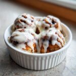 Four freshly baked small batch cinnamon rolls covered in thick white icing in a white ramekin.