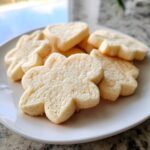 Close-up of several soft, un-iced vegan sugar cookies in flower and heart shapes piled on a white plate.