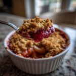 A spoonful of bright red, gooey rhubarb crisp filling and golden crumble topping being lifted from a white ramekin.