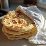 A warm stack of freshly made pita bread wrapped partially in a white kitchen towel.