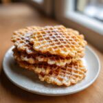 A stack of golden brown pizzelle cookies dusted generously with powdered sugar, resting on a light blue plate.