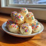 A mound of white, round sugar cookie truffles generously topped with colorful rainbow sprinkles on a white plate.
