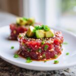 Close-up of two perfectly molded portions of vibrant red tuna tartare, topped with sliced avocado and green onions.