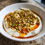 A close-up of a creamy whipped ricotta dip topped generously with chopped pistachios and drizzled with honey or chili oil.