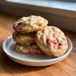 A stack of four soft, chewy white chocolate raspberry cookies featuring visible chunks of white chocolate and bright red raspberry pieces.