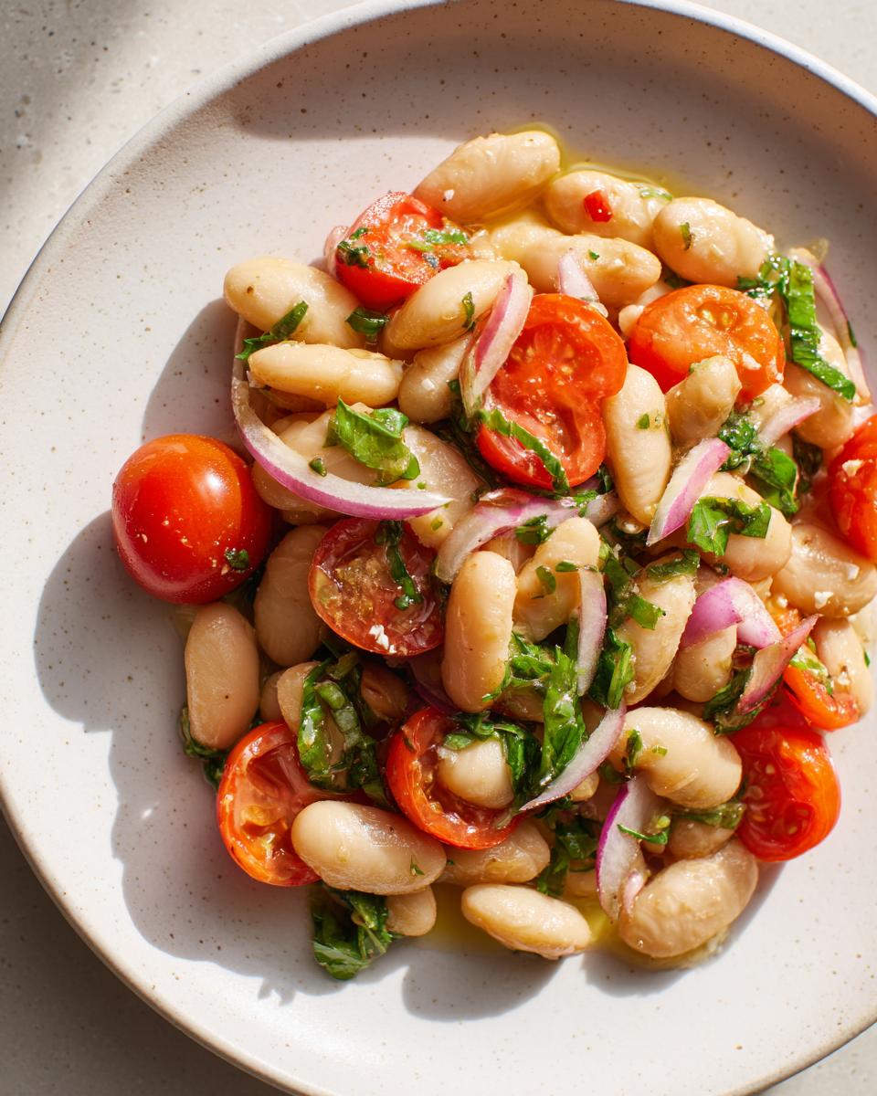 A close-up overhead shot of a bright white bean salad mixed with halved cherry tomatoes, red onion slices, and herbs.